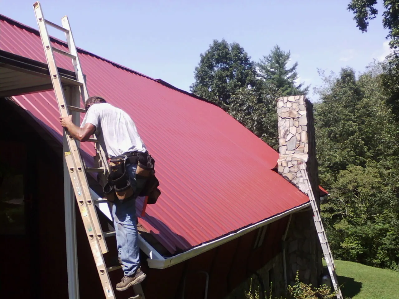 Roofer on a ladder beside a red metal roof and stone chimney — Soffit & Fascia Repair by Roofing Brothers in Greenbrier
